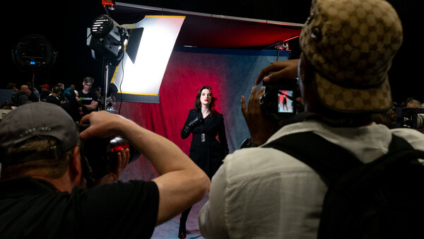 Audience members photograph a speaker on stage lit by professional studio lighting against a red and blue backdrop.