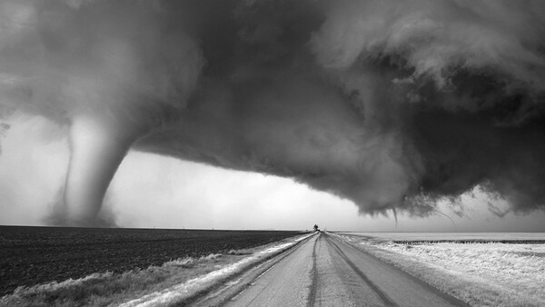 Tornado touching down across flat plains with a straight road leading toward the storm under dark clouds.