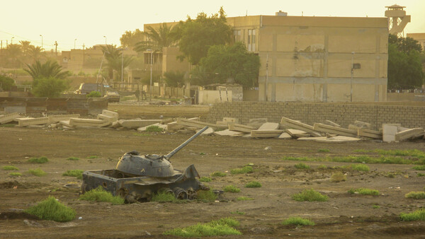 Abandoned tank turret in an overgrown field with deteriorating buildings and palm trees in the background.