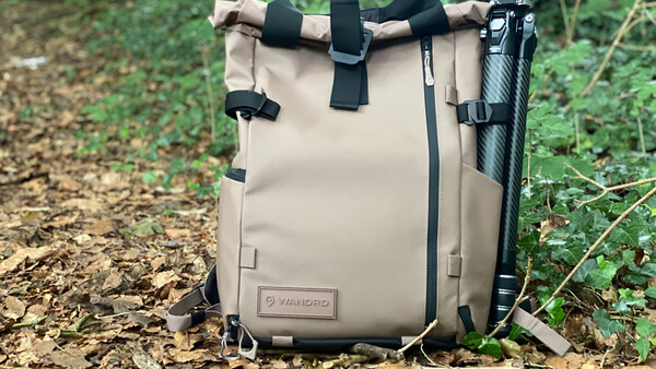 Tan canvas camera backpack with black straps and tripod attachment, photographed outdoors on forest ground.