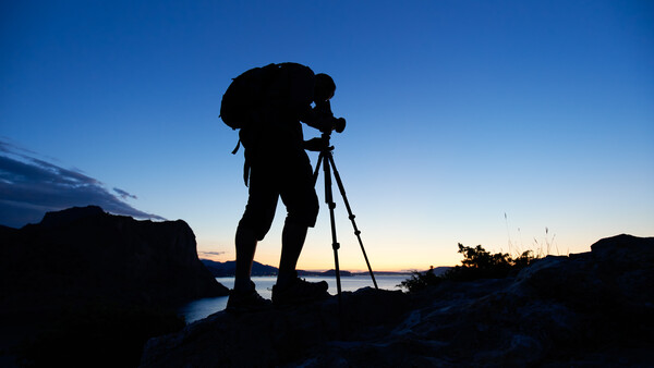 Silhouetted photographer with tripod on rocky coastline during twilight.