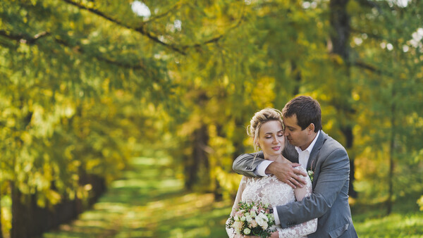 Groom embracing bride from behind in tree-lined garden path during golden hour.