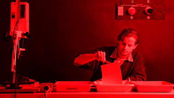 Photographer working in a red-lit darkroom, handling photographic paper under safelight conditions.