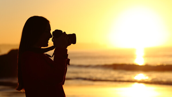 Silhouetted photographer holding a camera up to capture the sun setting over the ocean.