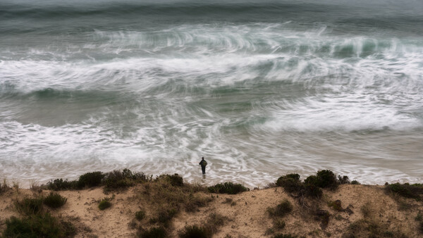 Solitary surfer standing on sandy beach with dune vegetation, facing large rolling waves under overcast sky.