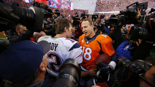 Football player in orange jersey surrounded by photographers and media at stadium field level.