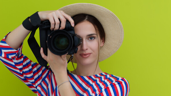 Woman in striped shirt and straw hat holding DSLR camera up to her face against yellow background.