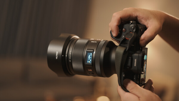 Hands holding a mirrorless camera with attached lens against a warm, blurred background.