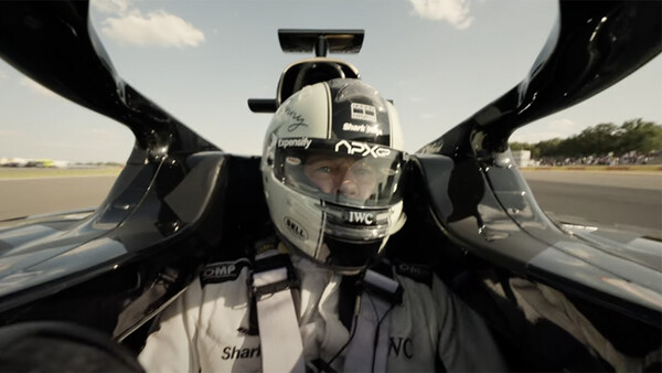 Driver in racing helmet and suit inside open-cockpit race car, captured from above on runway.