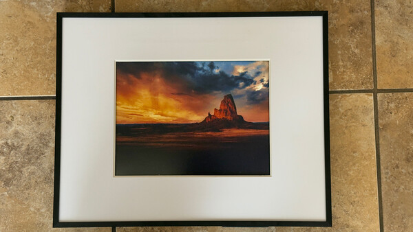 Framed photograph of a dramatic sunset over a desert rock formation with golden and orange sky.