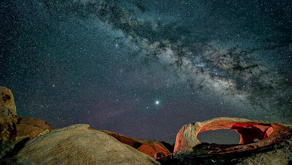 Milky Way galaxy stretching across night sky above desert rock formations with natural arch.