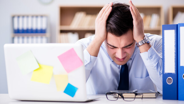 Stressed businessman at desk with head in hands, laptop covered in colorful sticky notes.