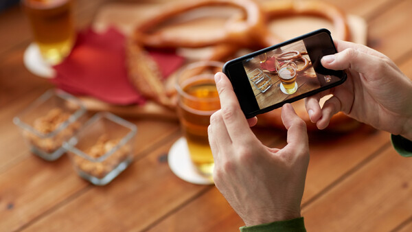 Person holding a smartphone capturing a flat lay composition of appetizers and drinks on a wooden table.