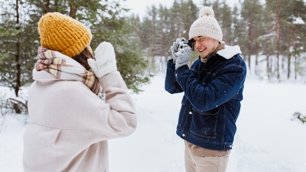 Two people playing in snow, one in a white jacket and yellow beanie, the other in a denim jacket and white beanie, with evergreen trees and snowy landscape in background.