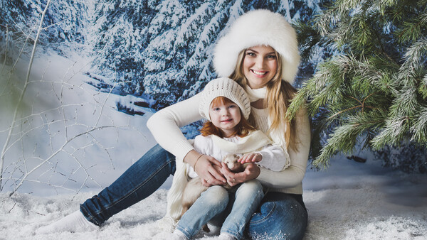 Woman and child sitting together in snow wearing matching cream-colored winter hats and sweaters.