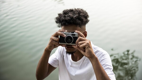 Person holding vintage rangefinder camera up to face, positioned by water with natural overcast lighting.