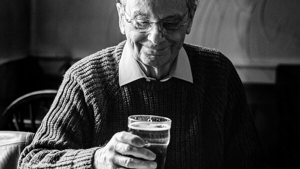 Older man wearing glasses and a cardigan holding a glass of water, looking down with a gentle expression.