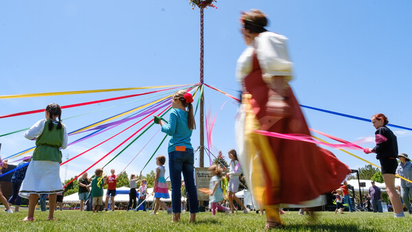 Children and adults holding colorful ribbons attached to a tall maypole during an outdoor spring celebration on a sunny day.