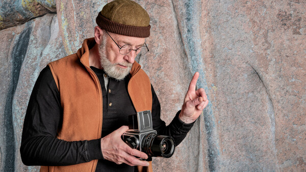 Older man with beard and glasses holding a vintage film camera against a textured stone wall.