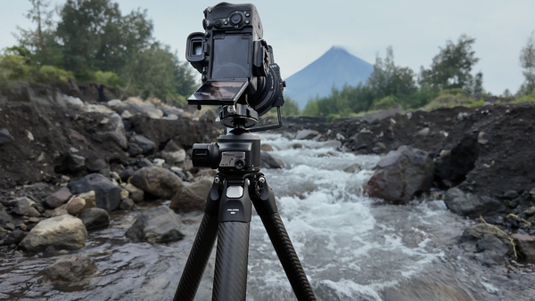 Professional camera mounted on tripod overlooking a rushing mountain river with volcanic peak in background.