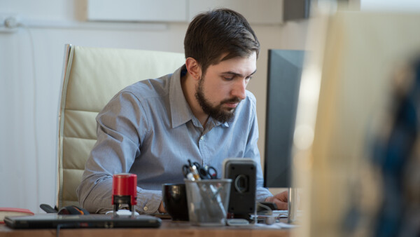 Male professional with beard seated at desk, focused on smartphone with studio lighting setup visible.