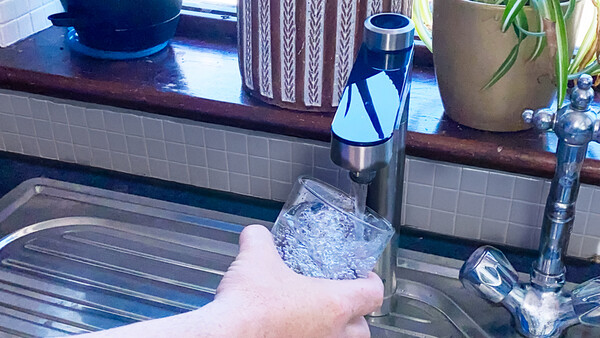 Hand holding a glass of water from a kitchen faucet with aerator visible.