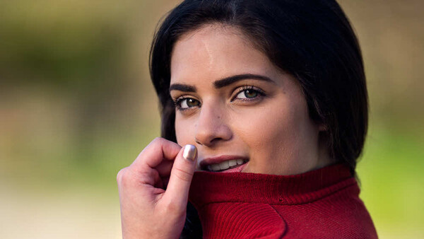 Woman with dark hair and red lipstick, wrapped in burgundy fabric, gazing at camera with hand near face.
