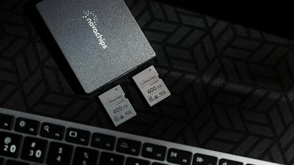 Two SD memory cards next to a dark gray storage case on a patterned surface beside a keyboard.