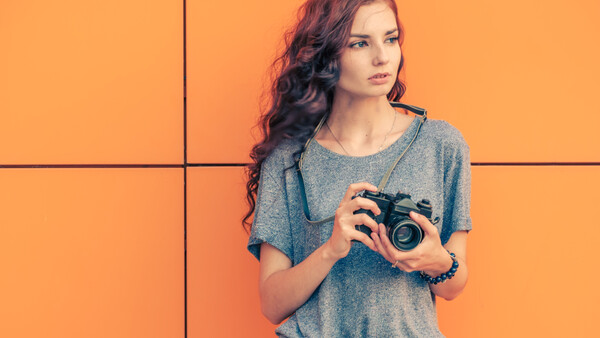 Woman with long wavy hair holding a vintage film camera against an orange background.