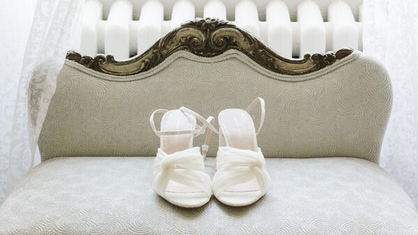 Pair of white infant shoes displayed on a cream-colored vintage chair with ornate wooden frame.