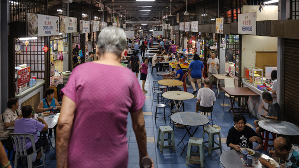 Woman in purple shirt walking through crowded indoor market or fair with vendor booths and tables.