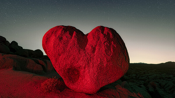 Large heart-shaped rock illuminated in vivid red against a dark desert landscape under a starry night sky.