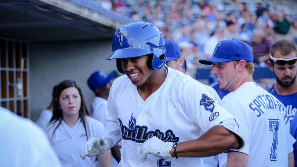 Baseball player in blue uniform smiling while being congratulated by teammates in dugout.