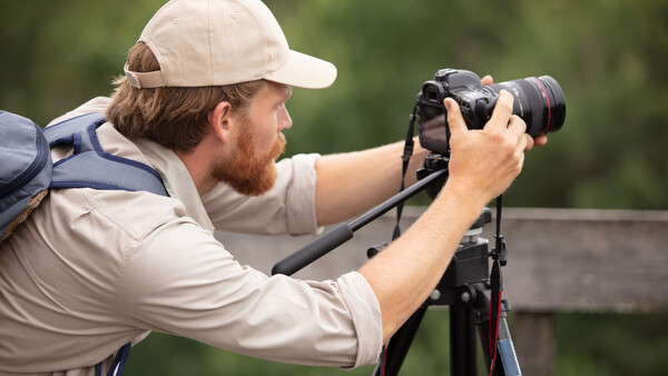 Photographer mounted on tripod with telephoto lens, wearing cap and neutral tones against blurred green background.