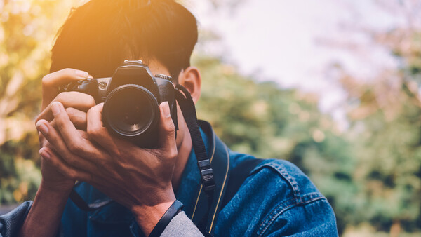 Photographer holding a DSLR camera up to their face, composing a shot outdoors in natural sunlight.