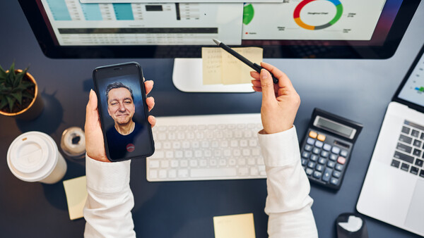 Person holding smartphone displaying a portrait photo while working at a desk with monitor and analytics dashboard.