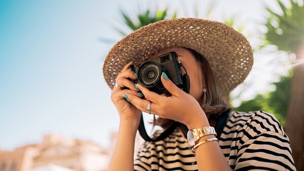 Woman in striped shirt and straw hat holding vintage camera to her face.
