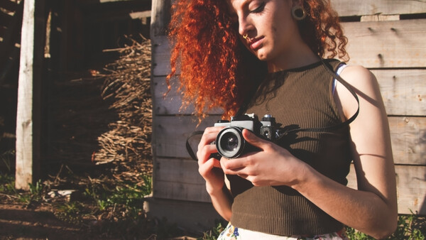 Woman with red curly hair holding vintage film camera outdoors.