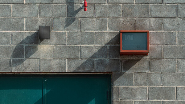 Minimalist urban wall with concrete blocks, red fire alarm box, and geometric shadows from sunlight.