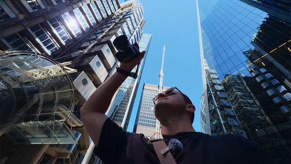 Photographer holding camera overhead while looking up at tall urban skyscrapers against blue sky.