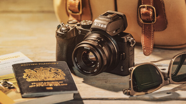 Nikon camera with lens surrounded by travel accessories including passport, sunglasses, and leather bag.