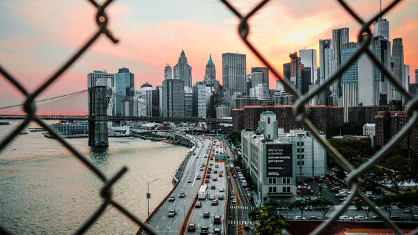 Manhattan skyline at sunset viewed through a chain-link fence from across the water.