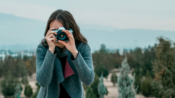 Woman holding a camera up to her face while standing outdoors with forested mountains in the background.