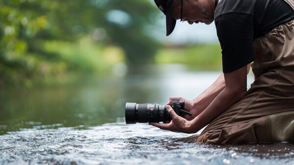 Photographer crouching in shallow water while holding a camera with telephoto lens.