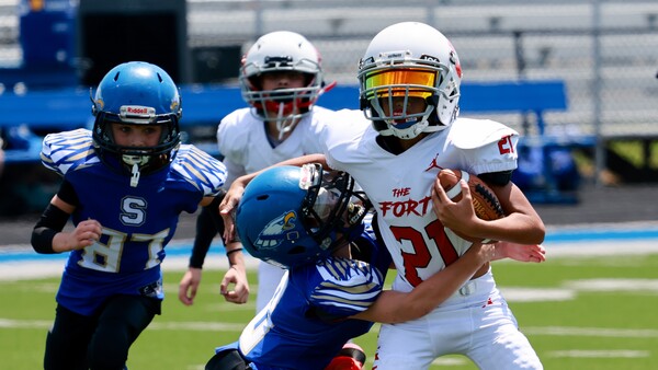 Youth football player in white uniform carrying ball while being tackled by defender in blue uniform.