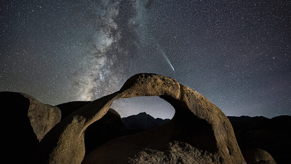 Milky Way galaxy arching over a natural rock arch formation under a starry night sky with a visible meteor.