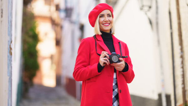 Woman in bright red coat and beret holding vintage camera on narrow European street.