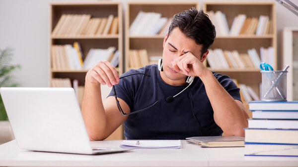 Man in dark shirt looking stressed while holding eyeglasses at desk with laptop and books.