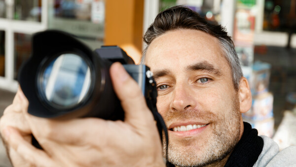 Male photographer holding a camera with a large telephoto lens, smiling at the camera.
