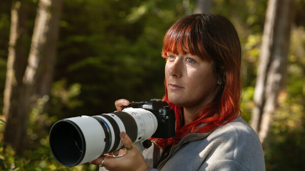 Female photographer with red hair holding a telephoto lens camera in a forest setting.